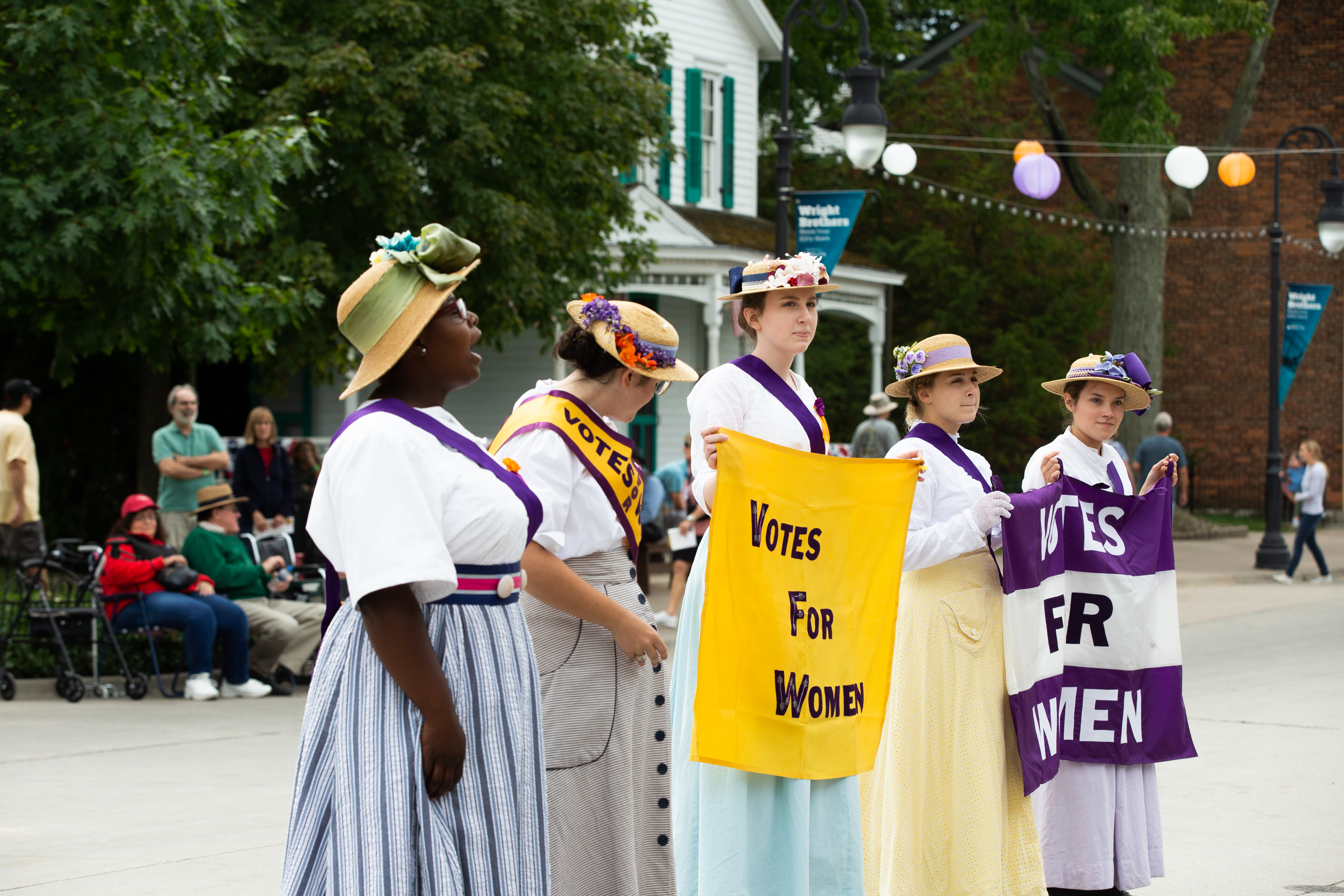 Presenters representing suffragettes Five women wearing sashes and holding signs related to women's suffrage