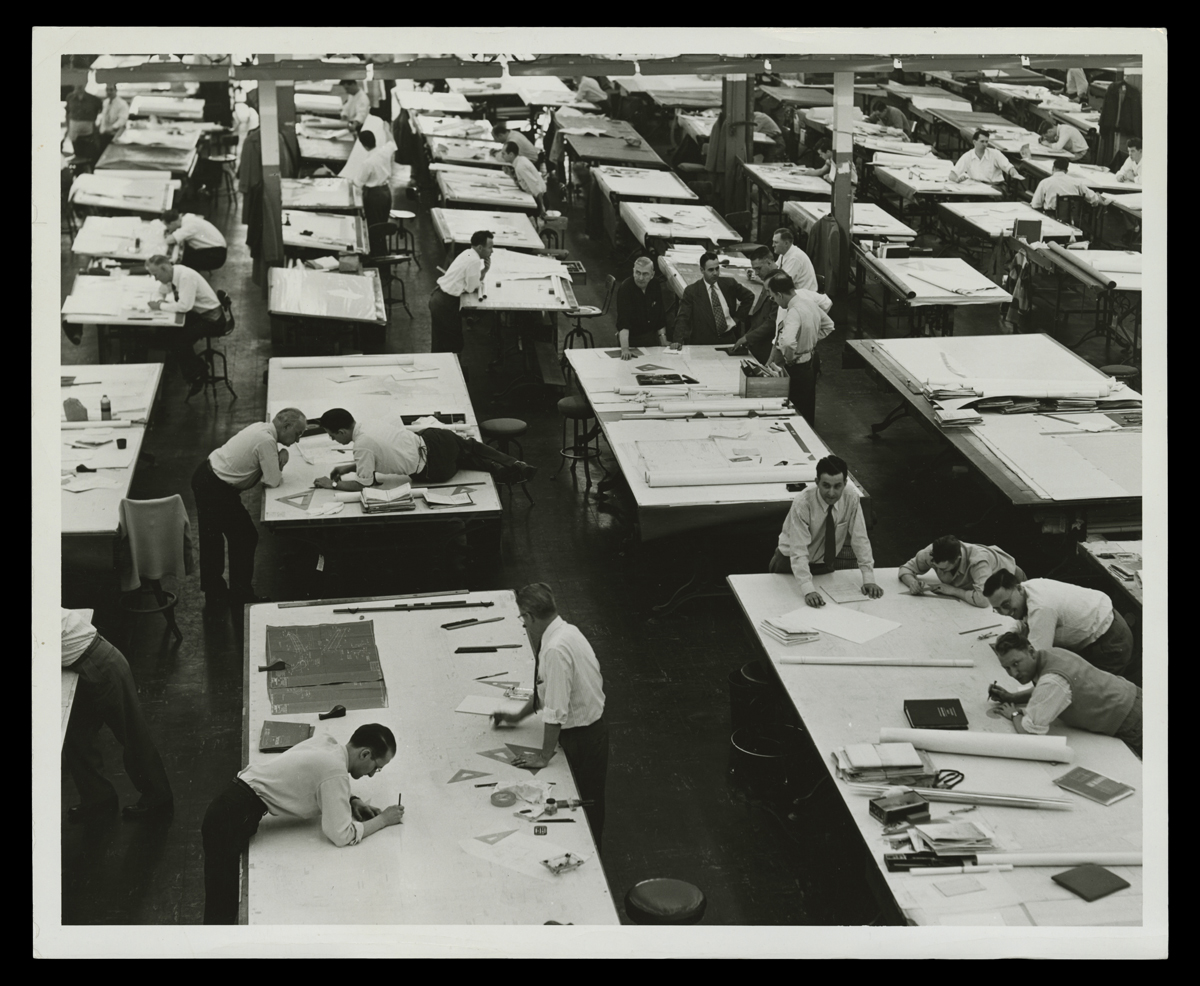 Large room full of tables, some of which have men standing near, leaning on, or lying on them with pencils or pens in hand
