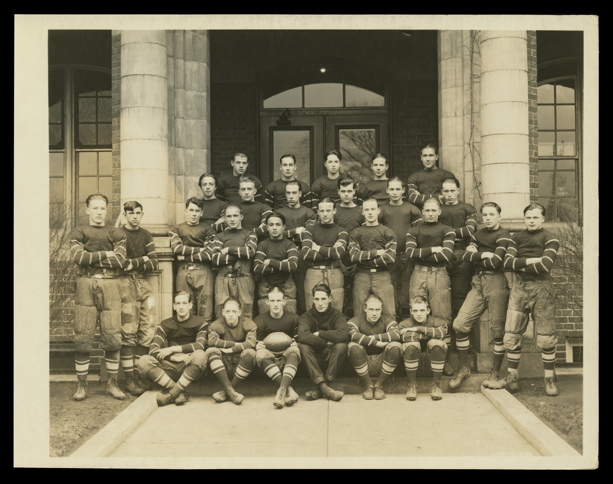 Henry Ford Trade School Football Team, 1923 Group of boys/young men wearing sports uniforms pose with arms crossed, seated and standing, in front of a building