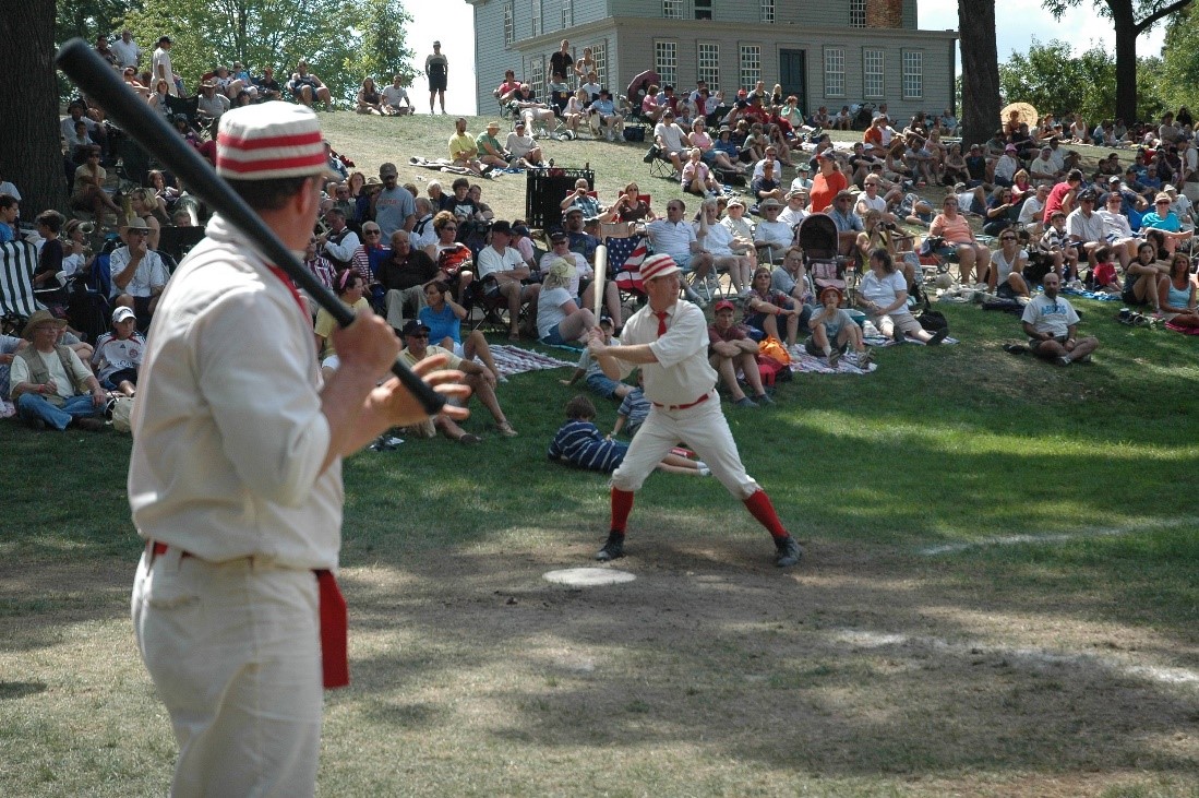 An historic base ball player holds a bat ready to swing as another stands nearby and a crowd looks on