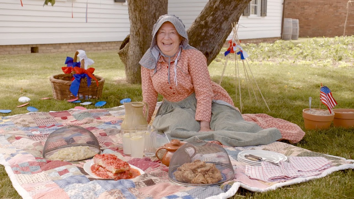 Cindy Melotti in 1876 centennial program at the Ford Home in Greenfield Village Woman in gingham dress, bonnet, and apron sits on a picnic blanket with food in front of a tree and white wooden house