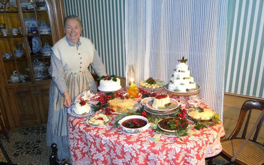 Cindy Melotti during Holiday Nights in Greenfield Village in the Ford Home Woman in dress and apron stands next to table with many holiday foods on it