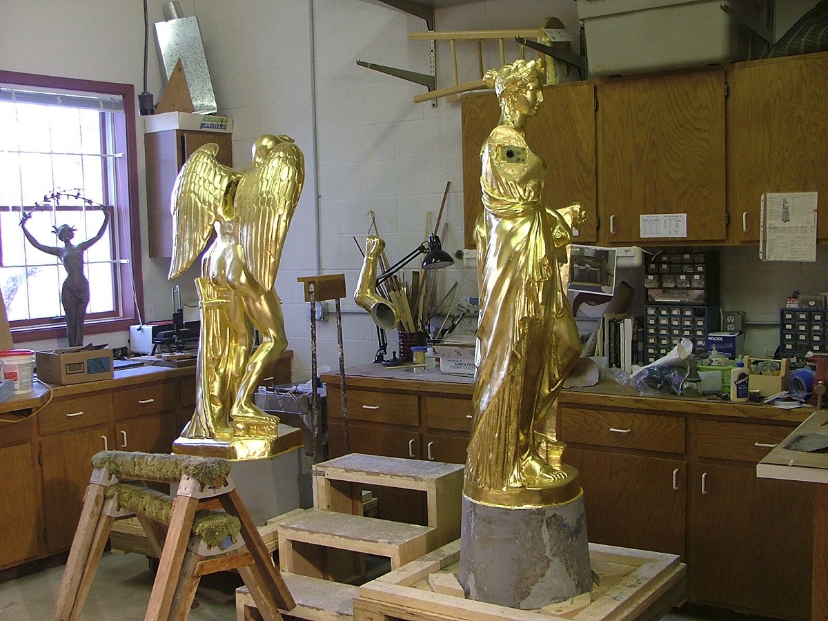 Two golden statues, one of a woman wearing flowing robes and one of a figure with wings, in a workroom