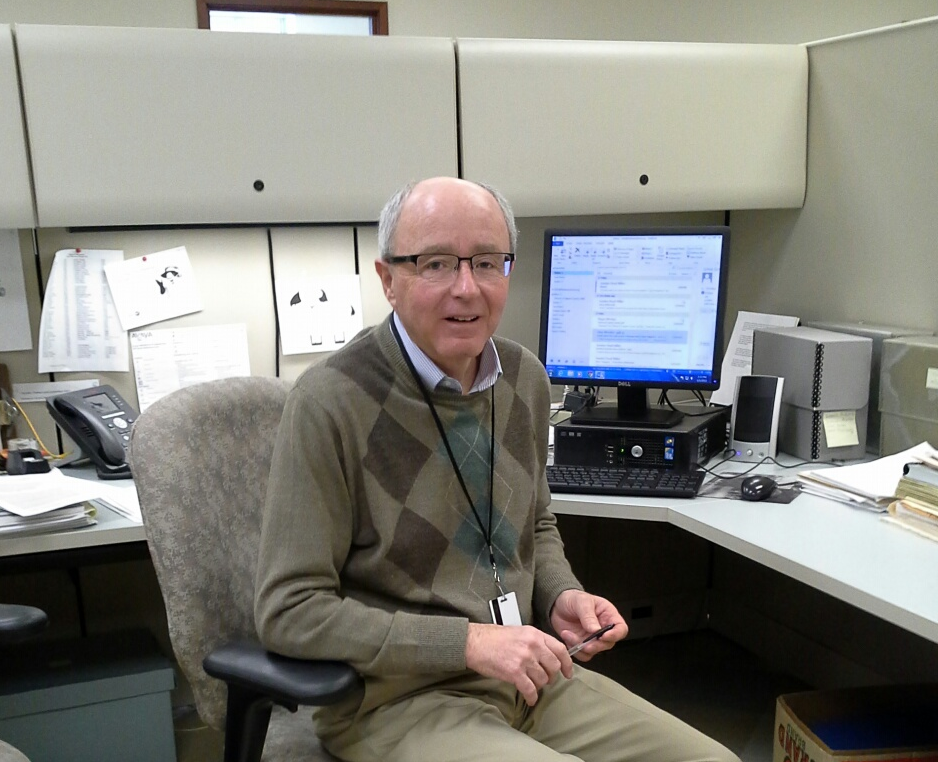 Curatorial Research Volunteer Gil Gallagher Man with glasses and white hair wearing an an argyle sweater and khaki pants sits at an office cubicle with a computer and silver archival boxes nearby