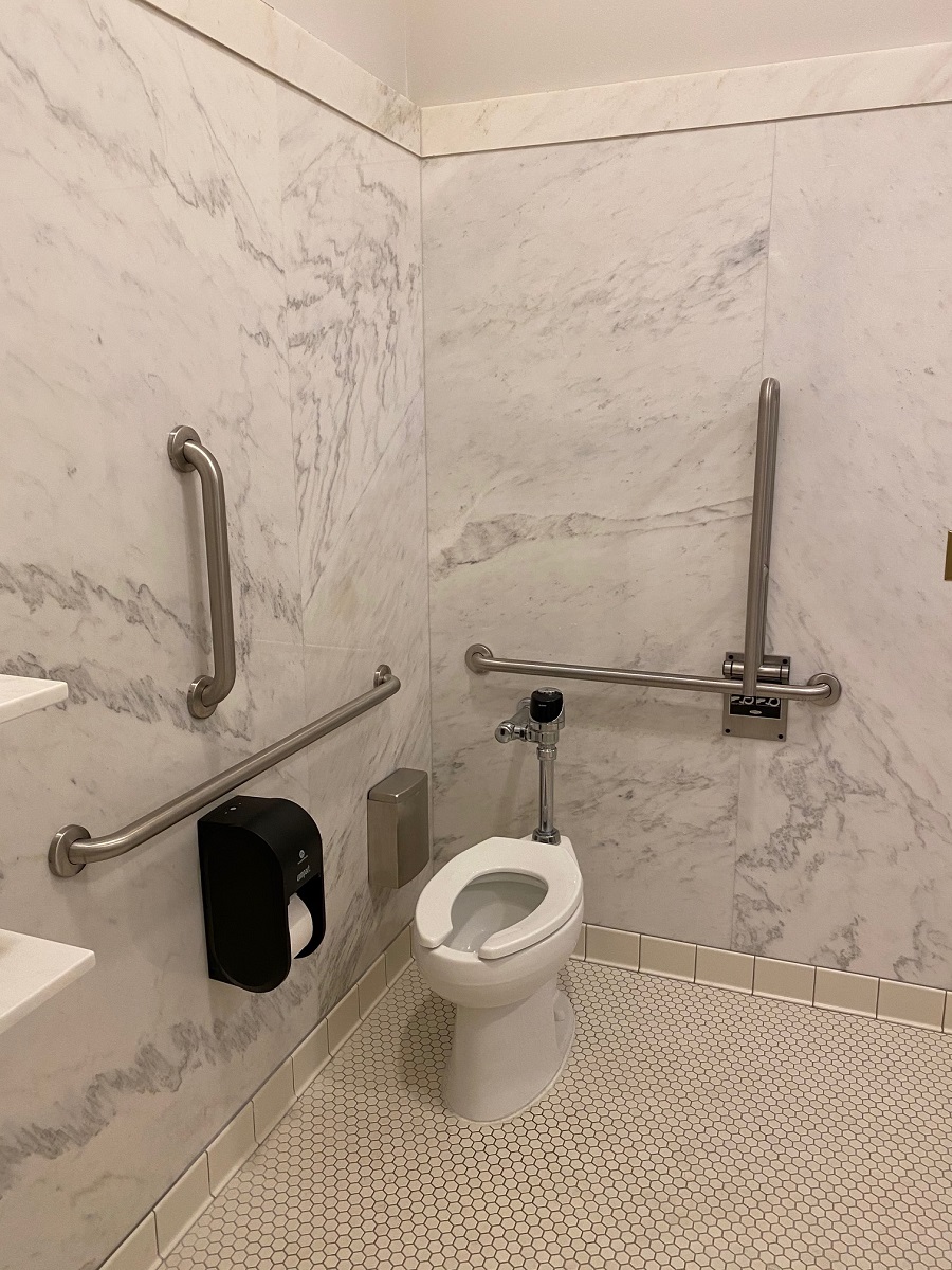 Toilet in one of our new accessible companion-care restrooms Toilet surrounded by vertical and horizontal grab bars in space with tile floor and marble walls