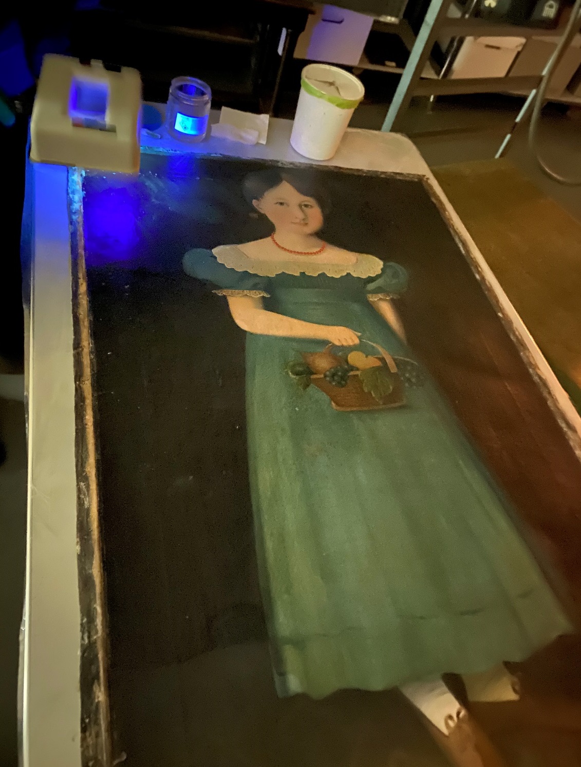 Painting of girl in blue dress, laid flat on table with blue light and tools at one end