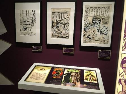 Three framed drawings with text and images on wall; additional text and images in lighted case below and to right
