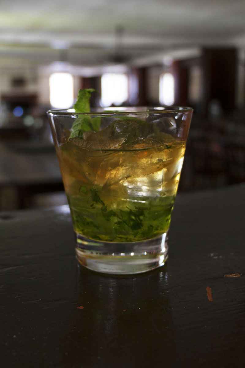 Drink in clear glass with mint leaf on top and muddled mint at bottom of glass; large dining room visible in background