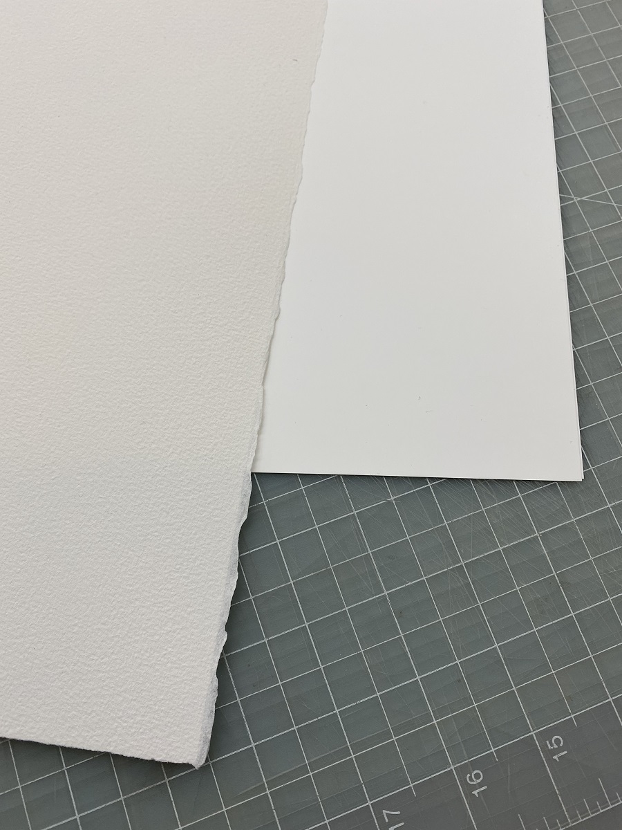Our paper options: A 74 lb. smooth, waterproof synthetic paper made of polypropylene, and a textured 90 lb. cotton fiber watercolor paper Two types of white paper lying overlapped on a grid background