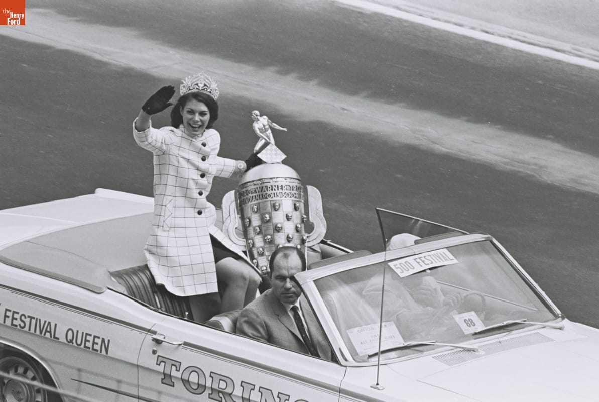 Festival Queen with the Borg-Warner Trophy at the 52nd Indianapolis 500, May 30, 1968 Woman wearing checked coat, tiara, and gloves and holding a very large trophy sits on the back of an open convertible and waves, with two men in the front seats
