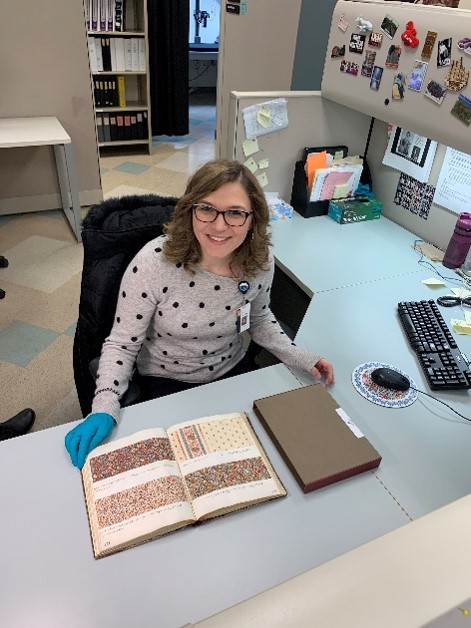 Project Cataloging Specialist Katrina Wioncek Cataloging a Sample Book from the American Textile History Museum Collection. Woman in office cubicle with book open on desk in front of her