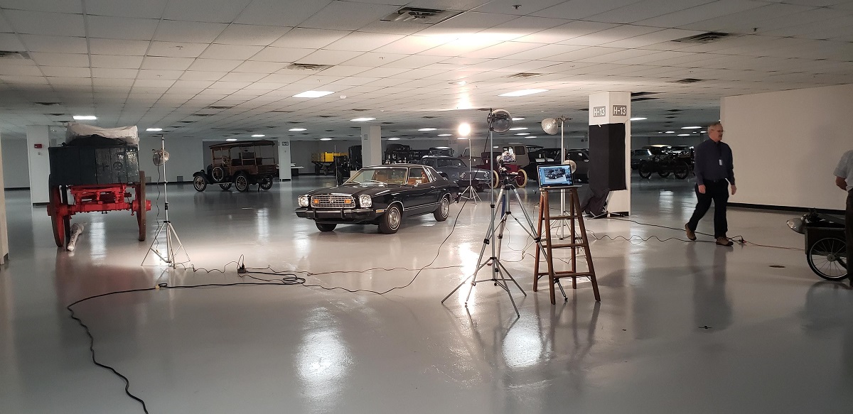 Photographing the Mustang II Black car surrounded by other vehicles in warehouse space; lights and camera on tripod pointed at it