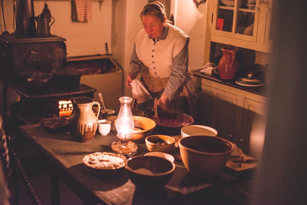 Historic presenter in the Ford Home during Holiday Nights in Greenfield Village Woman in candlelit kitchen with a variety of bowls and plates containing food on kitchen table