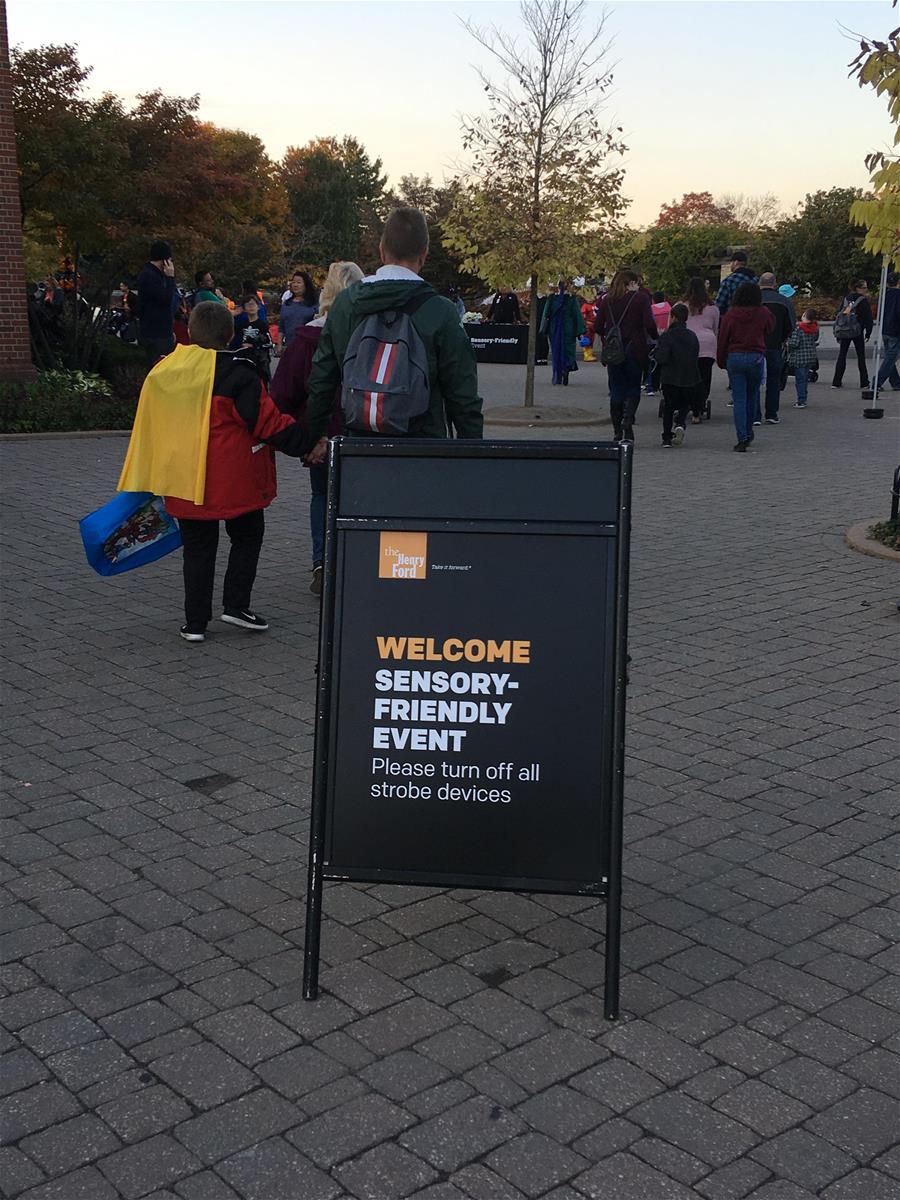 Sensory-Friendly Hallowe'en Entrance Sign Sign with text in foreground; plaza with many people and trees behind
