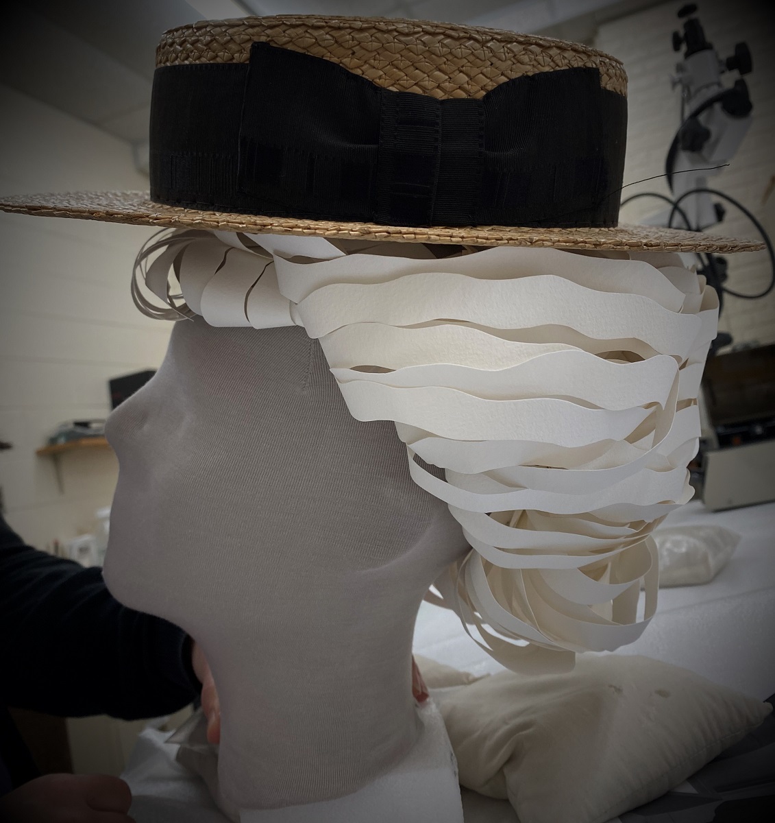 A handmade paper wig adorns the mannequin in our latest What We Wore exhibit Gray mannequin head with a white, cut-paper wig topped with a straw boater hat with wide black ribbon