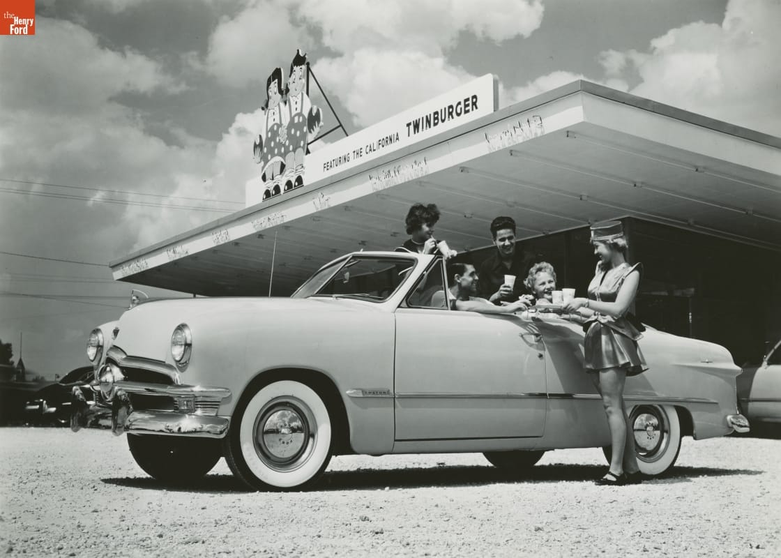 Ford Custom Convertible Club Coupe outside a Drive-In Restaurant, 1949 Black-and-white photo of a group of young adults in a convertible in front of a restaurant; a female carhop holds a tray by the car