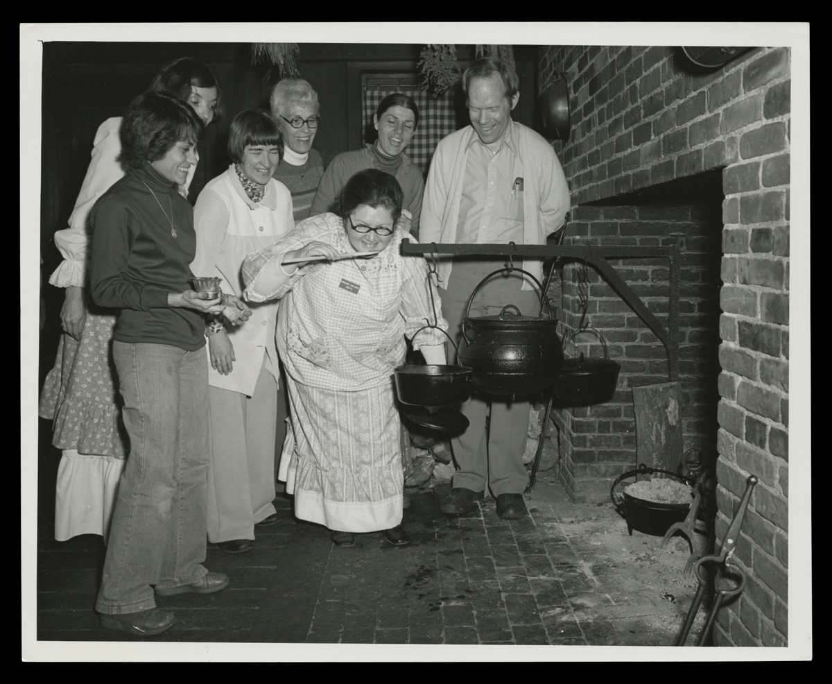 Colonial Cooking Class Held at Eagle Tavern in Greenfield Village, 1978 Group of people gathered around a fireplace and a cauldron on a swing arm