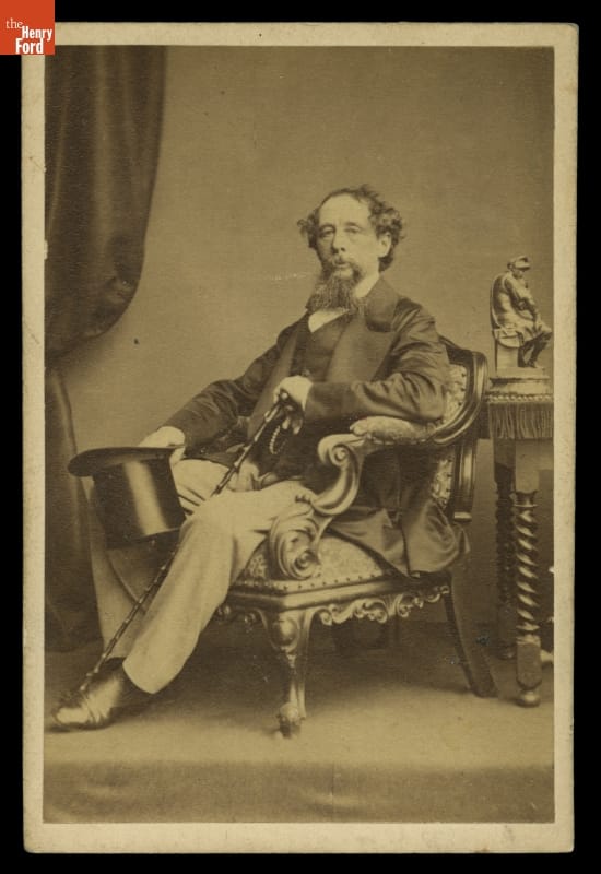 Man with beard and mustache seated in chair, holding cane and top hat, next to a table with a statue on it and drapery