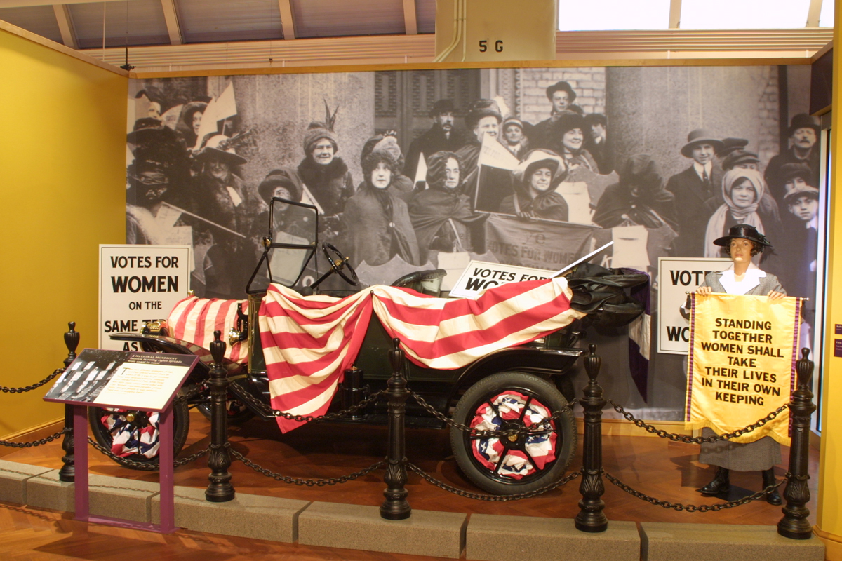 Model T covered in red and white striped bunting, in front of a photograph of women's suffrage advocates