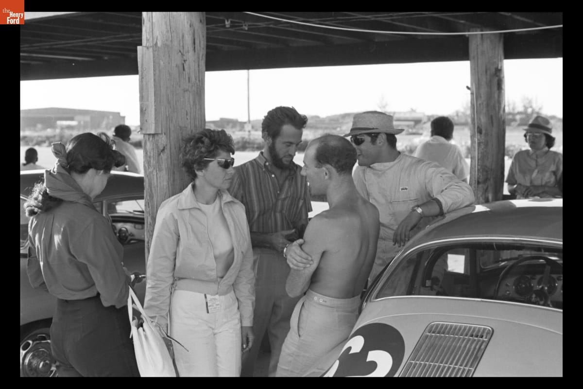 Denise McCluggage Talking with Stirling Moss at Bahamas Speed Weeks, November 27 - December 10, 1961 Woman leans against wooden post and talks to several other people, with additional people and cars nearby