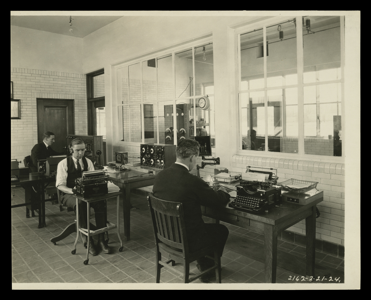 Men sit at desks with typewriters and equipment