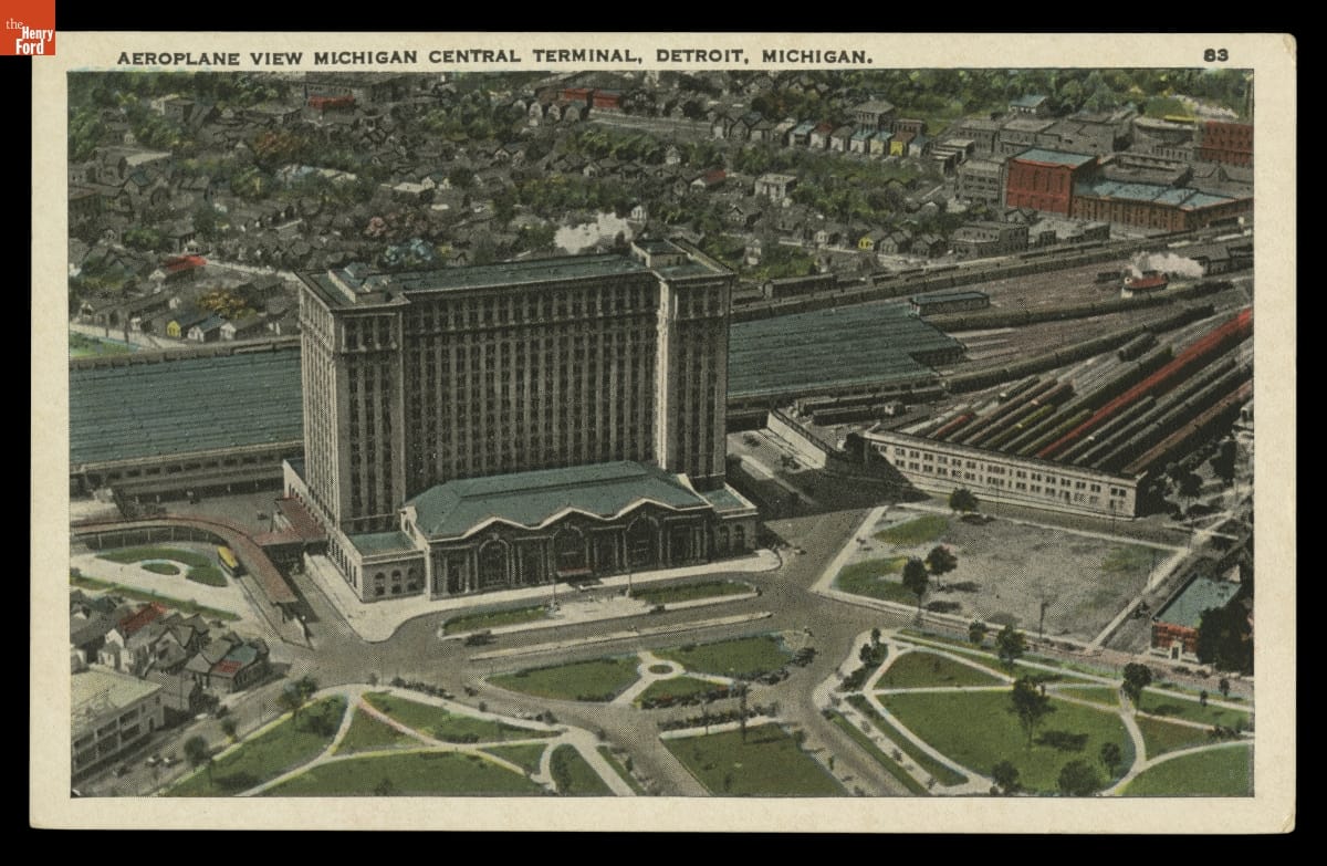 Aerial view of large building with railroad lines and trains behind and to one side of it; lawn and driveways in front and other buildings behind