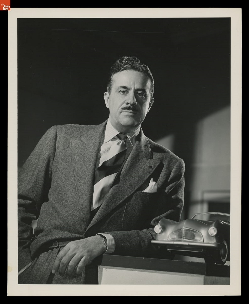 Portrait of Raymond Loewy, 1938-1949 Man with mustache wearing suit and striped tie leans on a surface holding a small car model