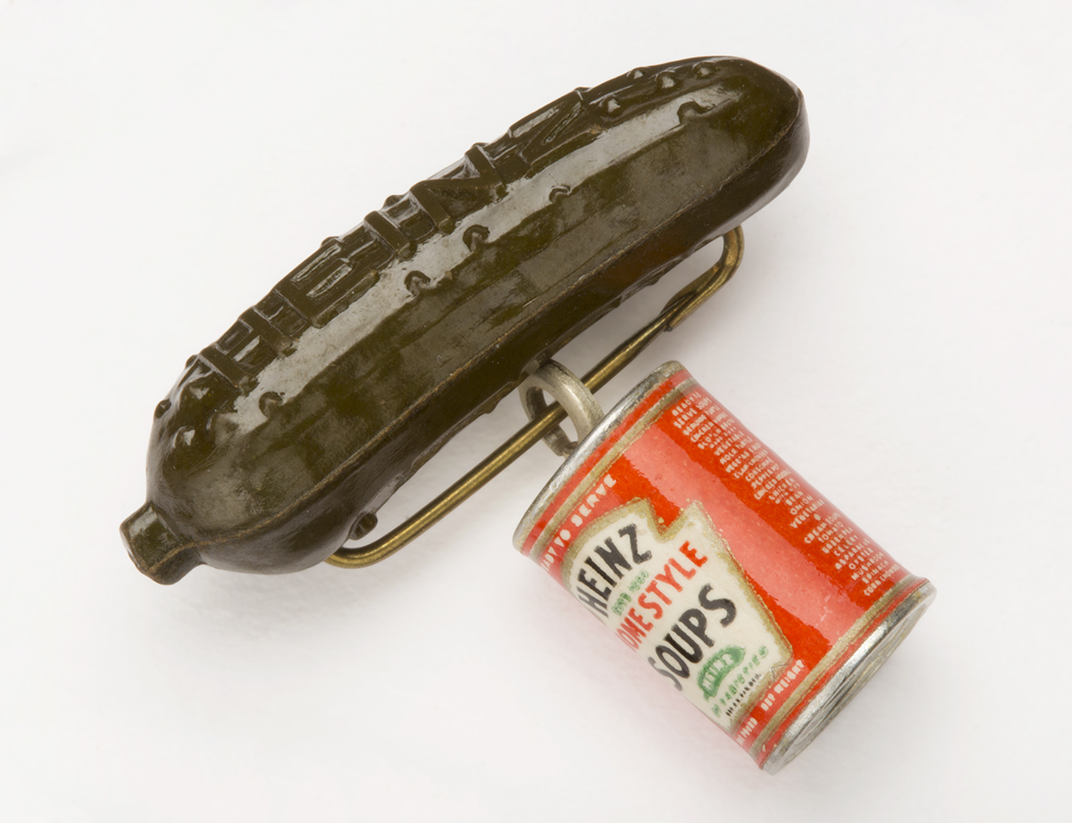 Pin in the shape of a green pickle with a red-and-white can of soup dangling from it; also contains text