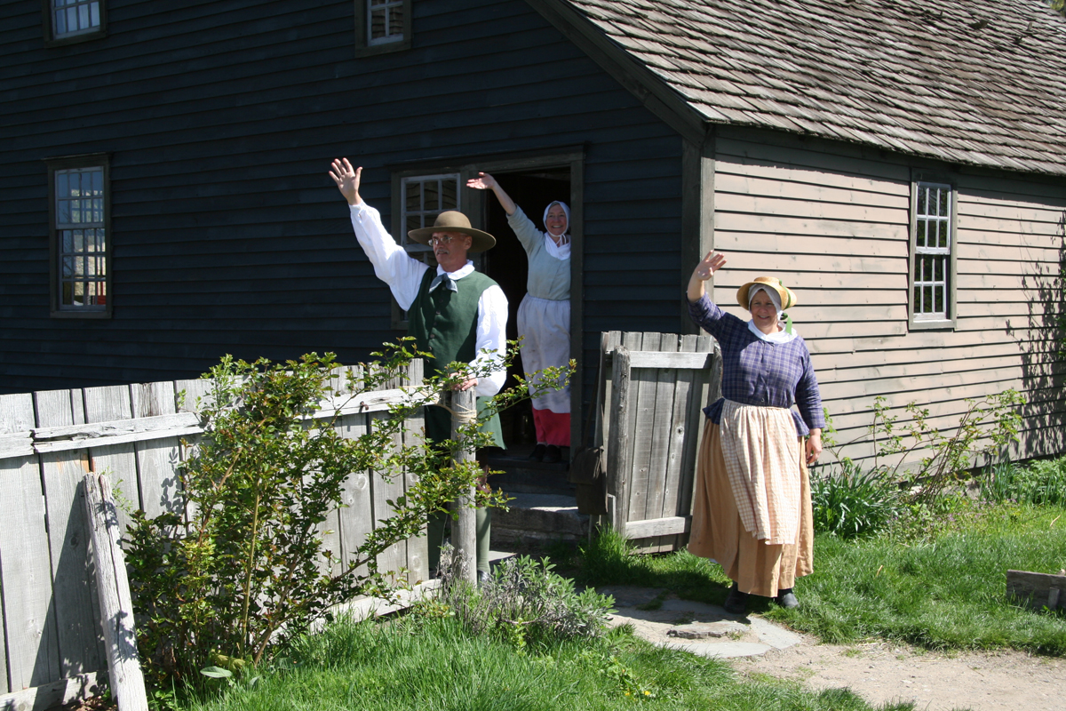 Three people in historic garb wave from the doorway and yard of a gray wooden building with a wooden fence