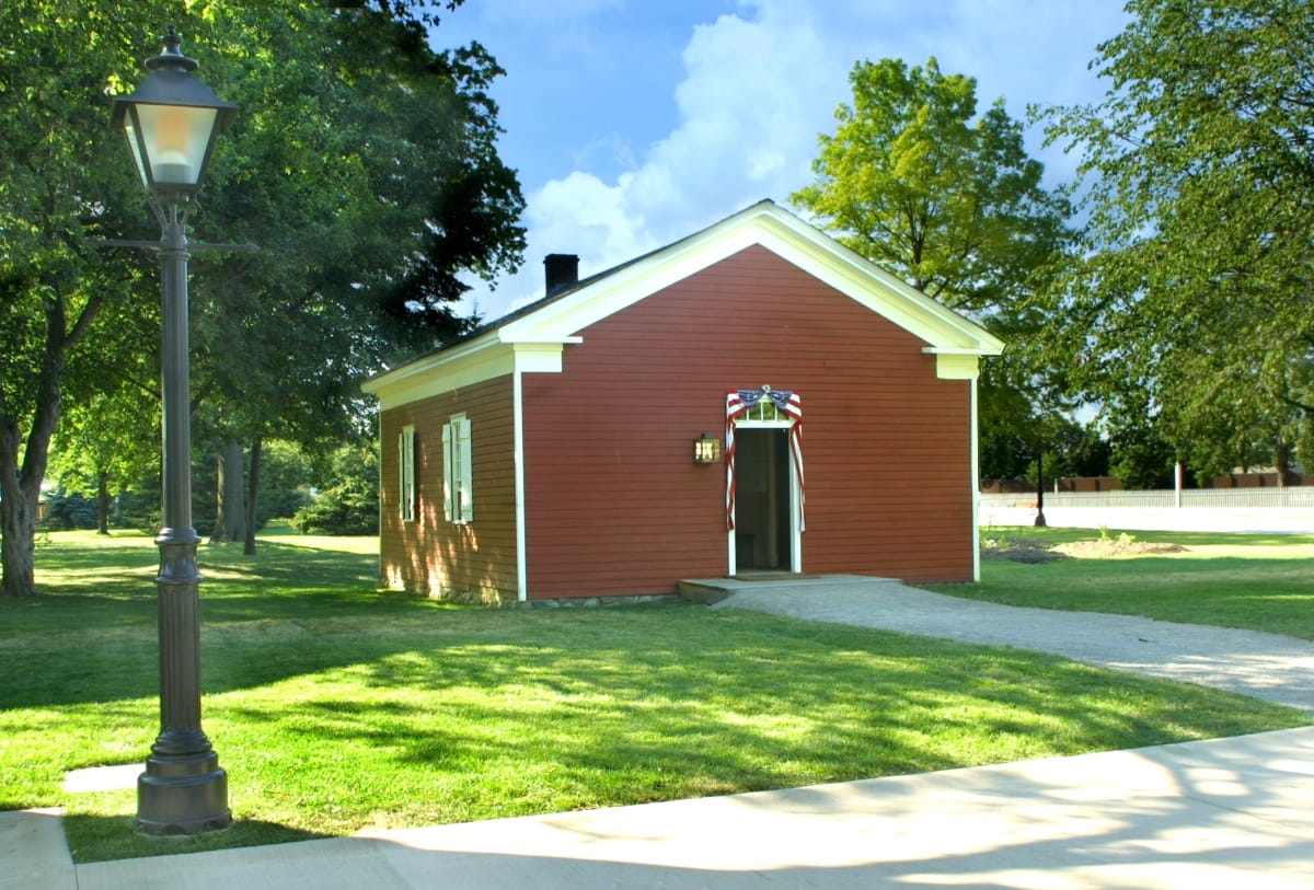 Small dark red wooden building on a large lawn with a streetlamp in front