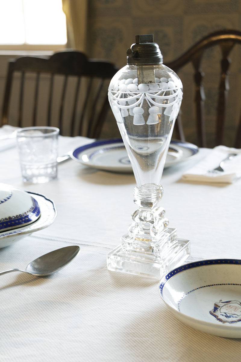 Noah Webster Home / whale oil lamp detail Clear glass lamp with etched pattern on tablecloth with dishes and silverware at place settings nearby