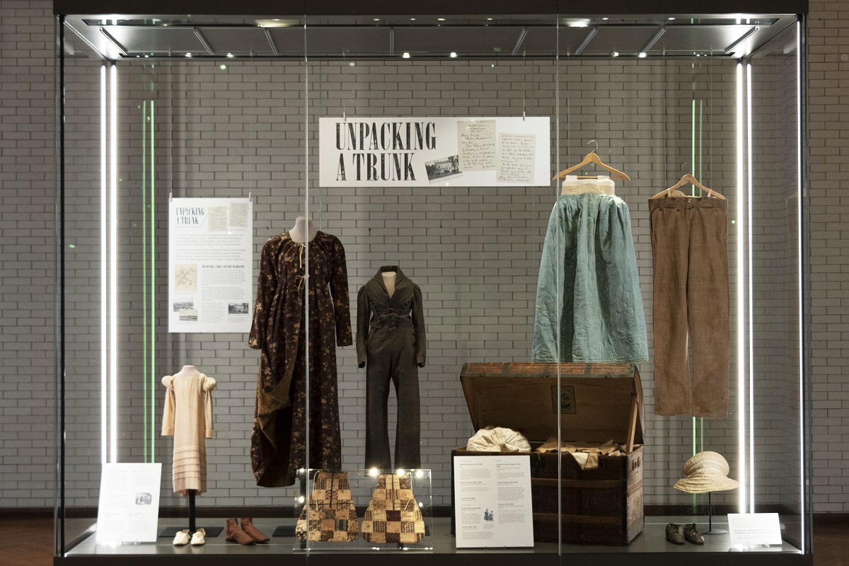 Large glass display case containing a wooden trunk and a number of garments and other pieces of apparel