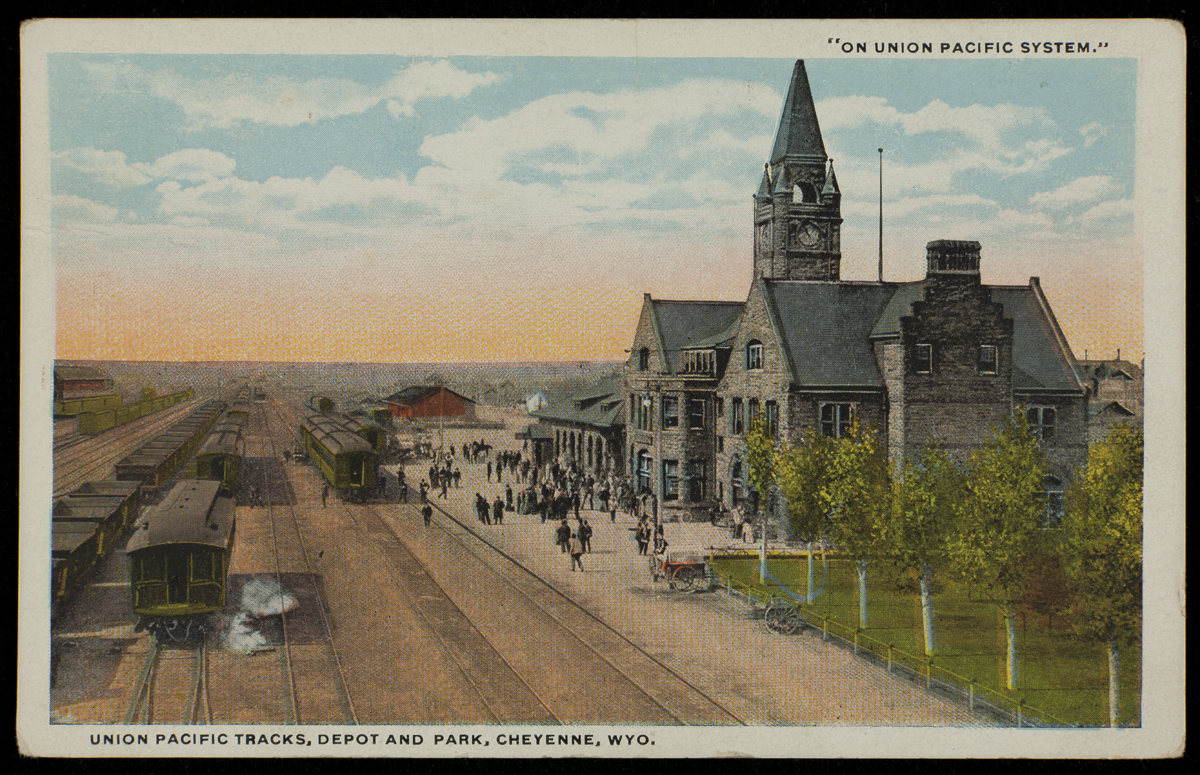 Postcard depicting large stone building with clocktower next to railroad tracks; people stand on platform between