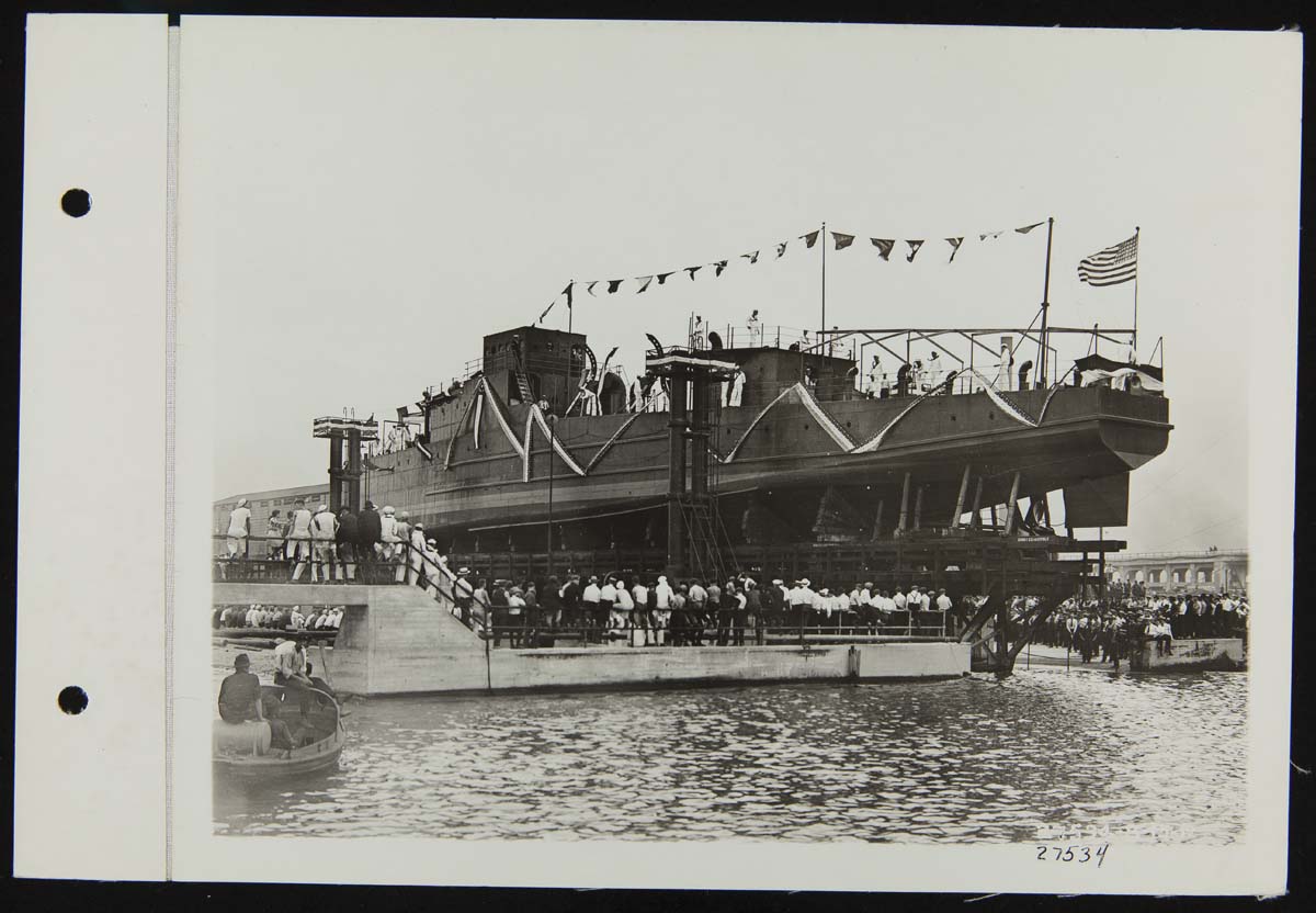 Long, narrow boat decorated with flags and bunting on elevated trestle above water, as a crowd looks on