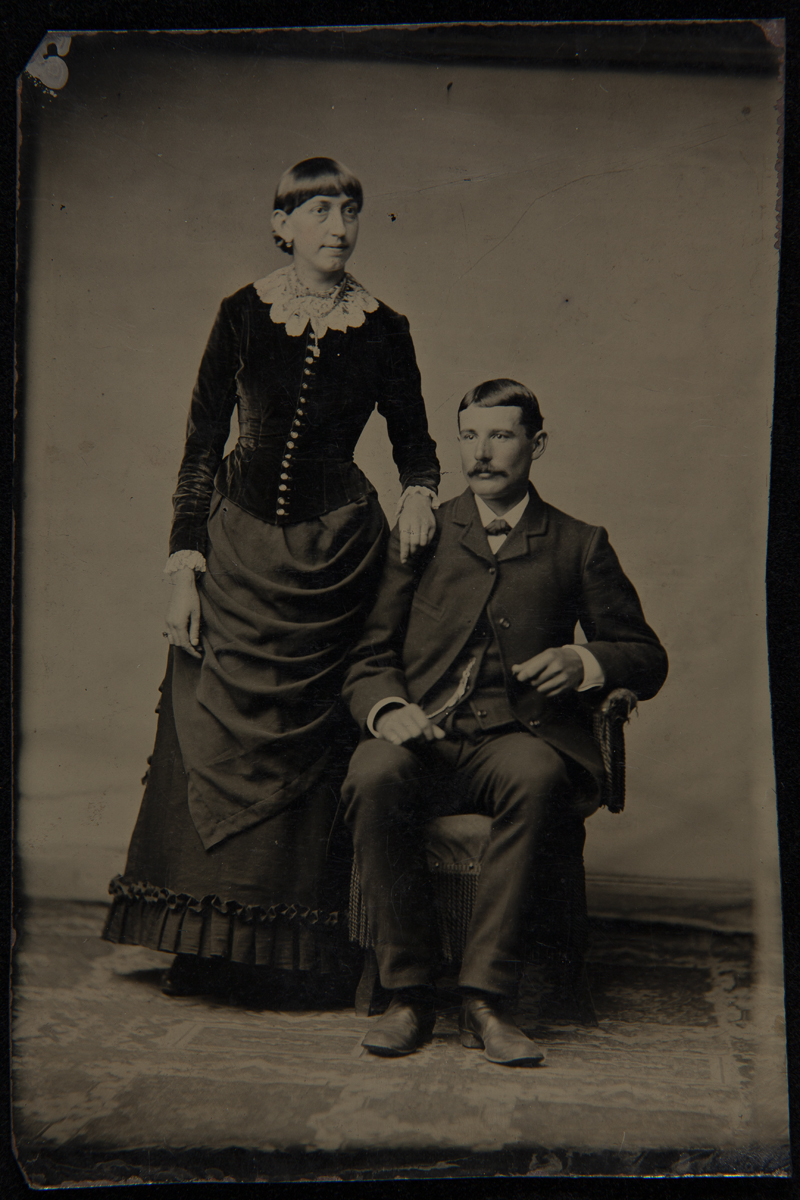 Wife with Lace Collar and Cuffs and Her Seated Husband, circa 1885 Woman with bangs wearing elaborate ensemble, standing with hand on shoulder of seated man wearing a suit