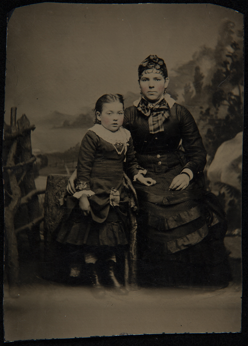 Mother and Daughter in Front of a Painted Landscape Backdrop, circa 1885 Seated woman and standing little girl, both in elaborate dresses, in front of a painted backdrop with logs as props