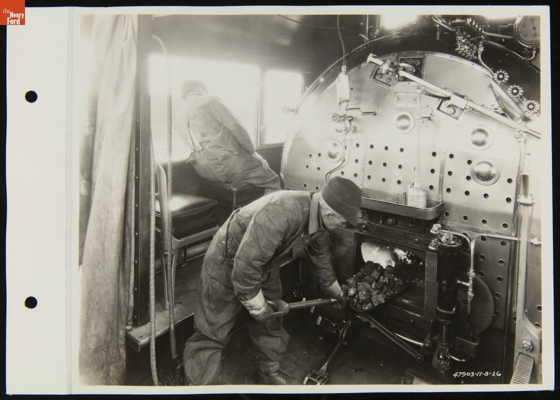 Railroad Fireman Shoveling Coal inside Steam Locomotive, Detroit, Toledo & Ironton Railroad, November 1926 Black-and-white image of two men, one shoveling coal into a large metal furnace and the other leaning out a window