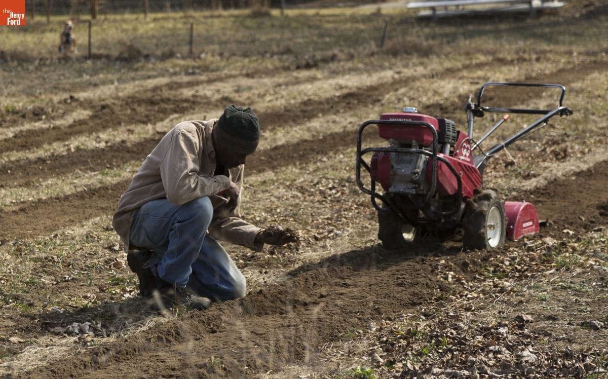 A Black man kneels in a field, examining soil, next to a tiller