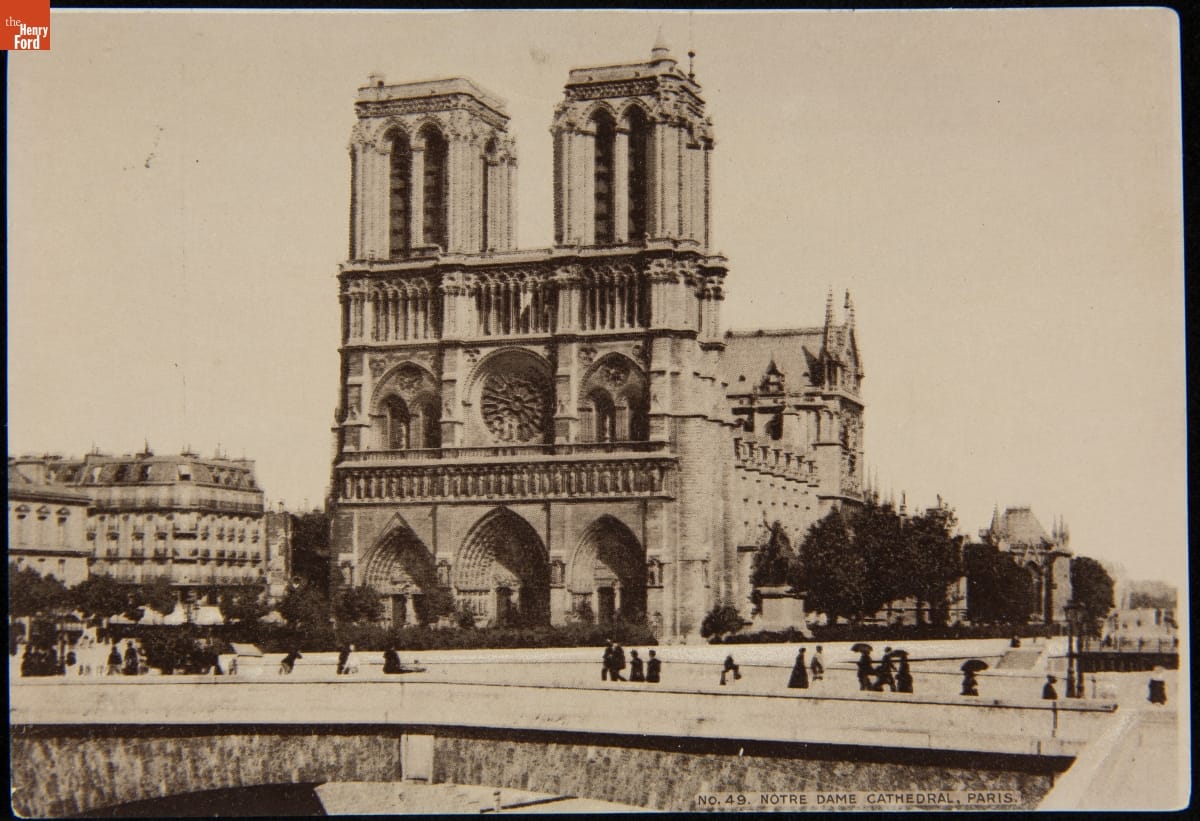 Black-and-white photo of gothic cathedral; people passing on roadway in front and other buildings visible to the side