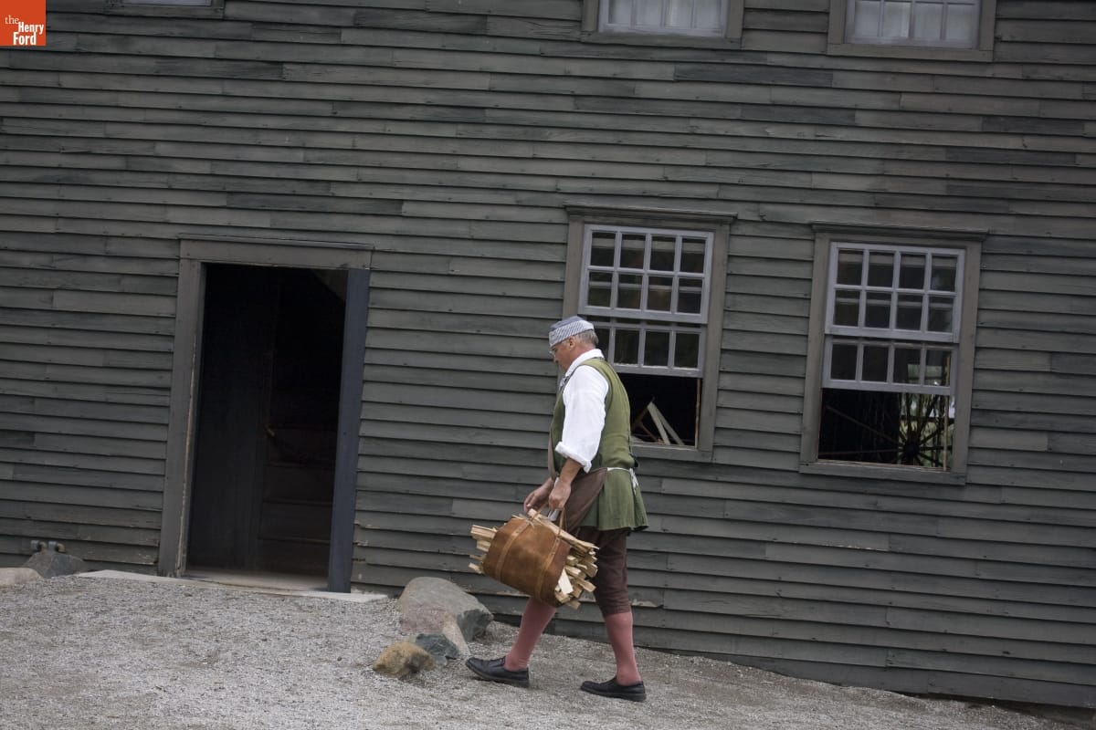 Man wearing historic clothing walks past simple gray wooden house