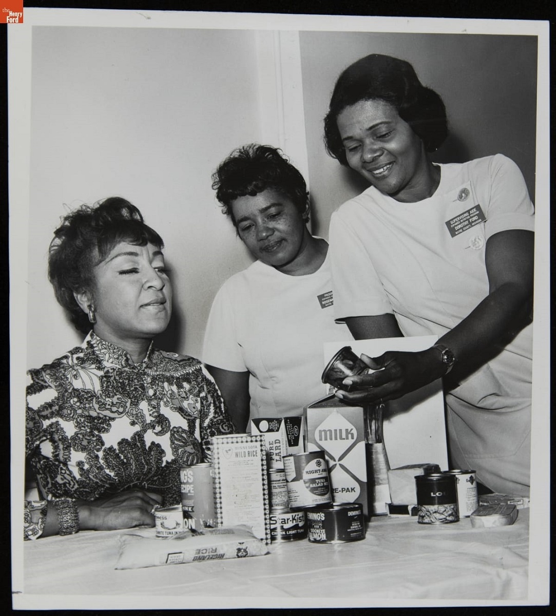 Three Black women sit and stand around a table that holds food packages