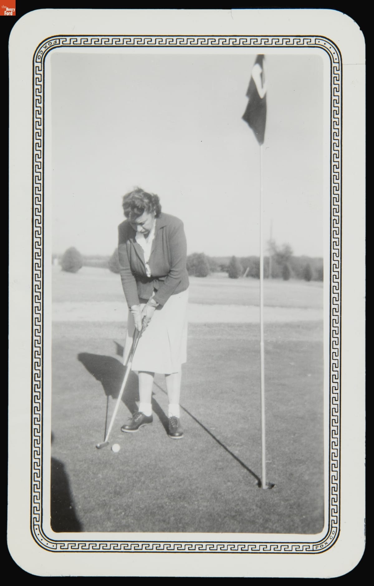 Woman Putting on a Golf Course, 1947 Woman in skirt and jacket bends over to putt a golf ball with hole marked with flag nearby