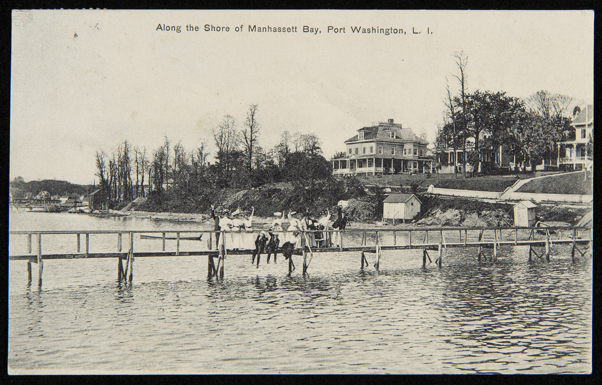 People wave from long, wooden dock over a body of water with houses visible along shoreline behind them; also contains text