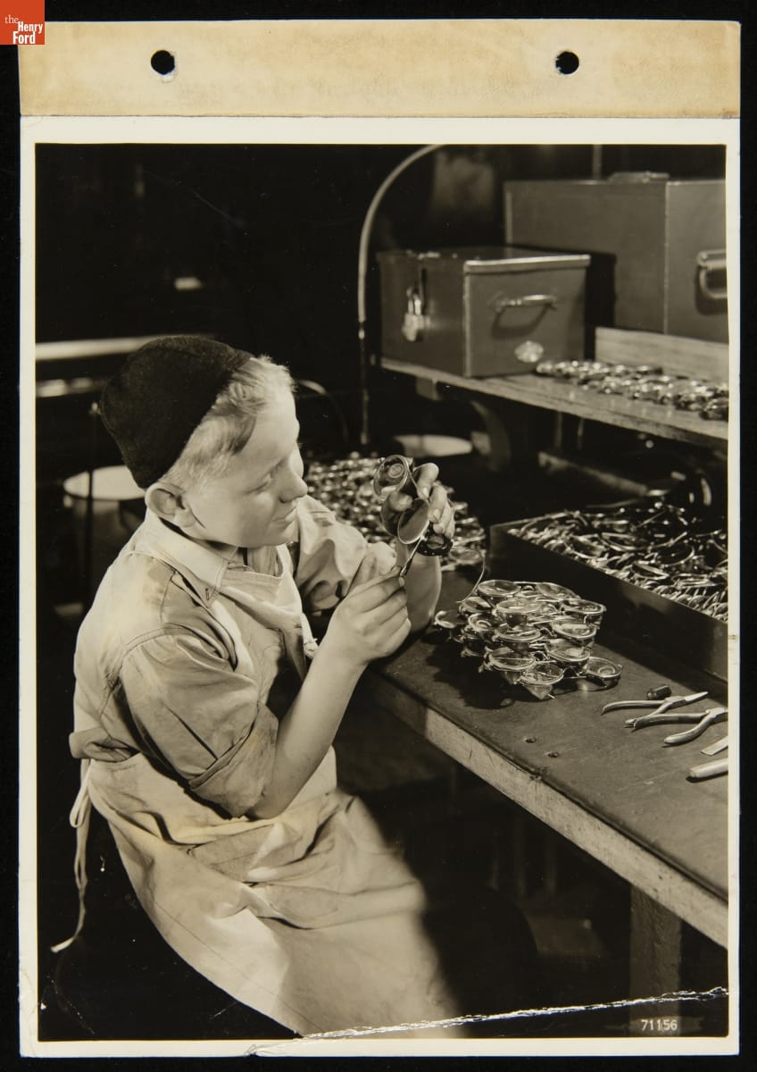 Henry Ford Trade School Student Repairing a Pair of Goggles, December 5, 1938 Boy wearing apron and cap holds tool to a pair of goggles; a table nearby holds many additional pairs of goggles