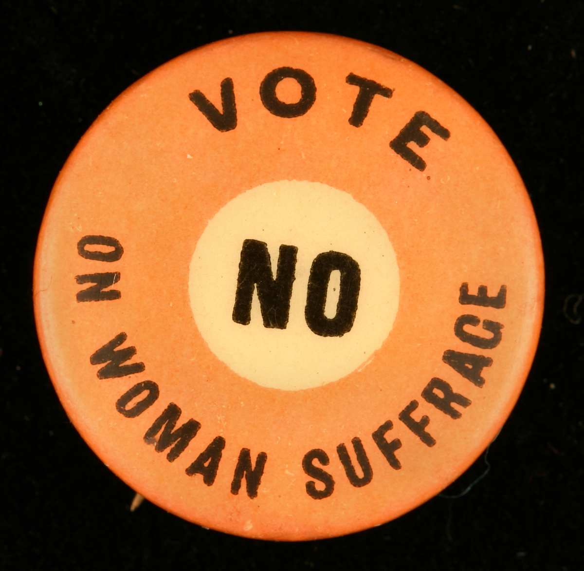 Round orange button with yellow circle in center, with printed text reading “Vote no on woman suffrage”