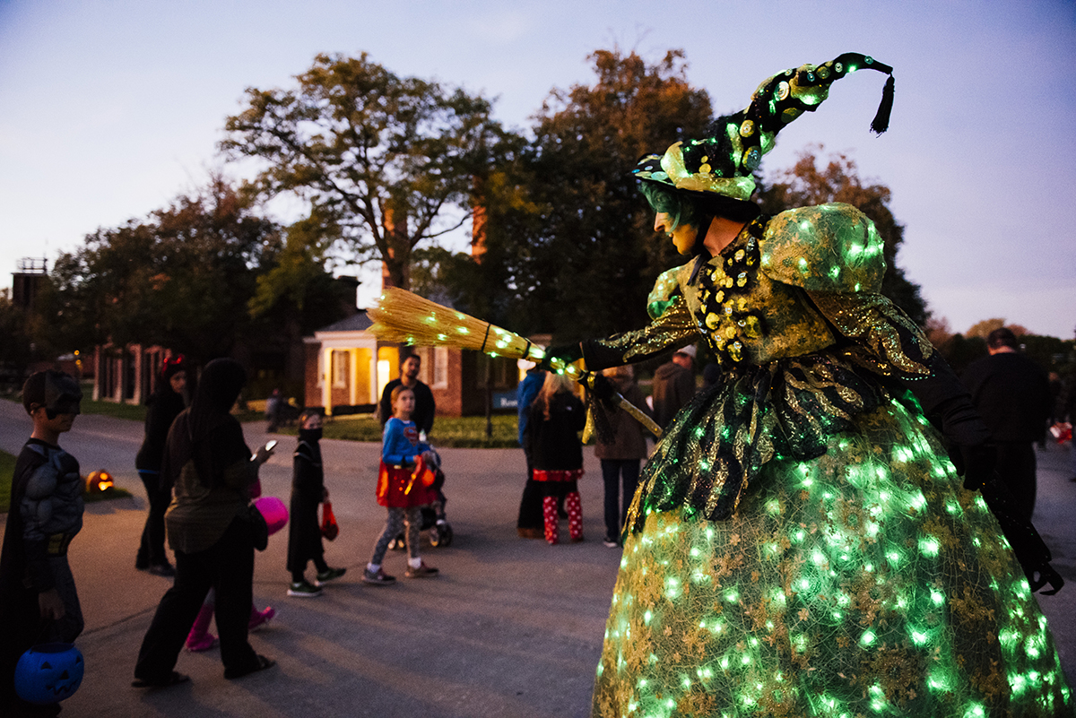 Hallowe'en in Greenfield Village, 2017 Woman in witch's costume decorated with green lights holds out a broom as children walk by